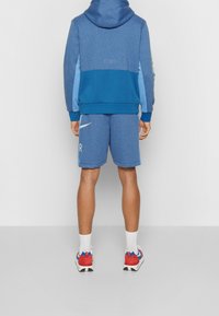 Blå hoodie med en ljusblå panel, som matchas med blå shorts med en vit logotyp. Vita strumpor och röda sportskor fulländar stilen.