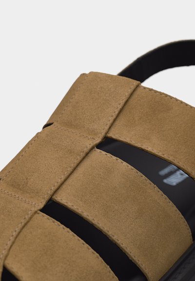 Sandale en daim marron avec des larges brides et des coutures visibles, dotée d'une semelle noire et d'un design à bout ouvert.