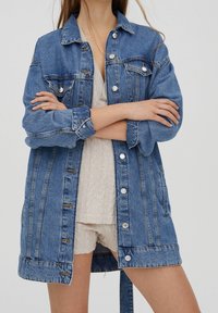 Veste en denim bleu avec ouverture boutonnée, deux poches poitrine et manches retroussées. Présente une coupe légèrement ample et des fentes latérales à l'ourlet.