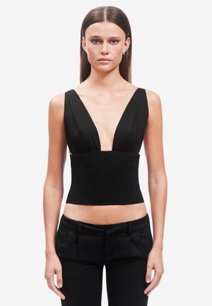 Frau mit glattem Haar, die ein ärmelloses schwarzes Crop-Top mit tiefem V-Ausschnitt und schwarze Hosen trägt, steht vor einem weißen Hintergrund.