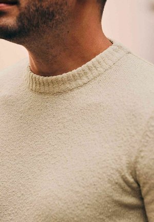 Man draagt een beige gestructureerde trui met ronde hals, waarbij de nek en het onderste gedeelte van het gezicht met lichte baardstoppel zichtbaar zijn tegen een effen achtergrond.