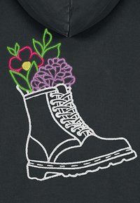 Fekete kapucnis pulóver, amelyen fehér hímzett mintázat látható: egy csizma tele színes virágokkal és levelekkel, rózsaszín, lila és zöld árnyalatokkal kiemelve.