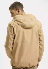 Beżowa, lekka kurtka z kapturem, z elastycznymi mankietami i gładką teksturą, zaprojektowana do noszenia na co dzień. Brak widocznego logo z tyłu.