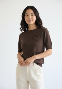 Brązowy sweter z krótkim rękawem, wykonany na drutach, z teksturowanymi kształtami serca na przodzie. W połączeniu z jasnymi spodniami.