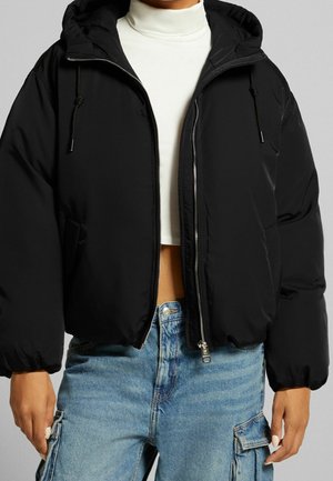Persoon met een zwarte donzen jas met capuchon, een witte cropped coltrui en blauwe denim cargobroek, staand tegen een effen achtergrond.