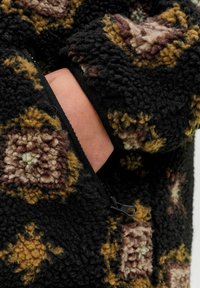 Crna flis jakna s teksturiranim završetkom, koja sadrži geometrijski uzorak u nijansama smeđe i zelene. Uključuje džep sa zatvaračem.
