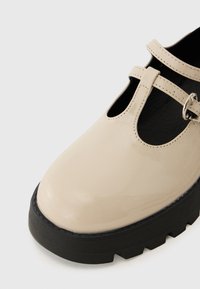 Zapato T-bar de charol beige con punta redonda, hebilla ajustable y una suela de goma negra gruesa con un patrón texturizado.