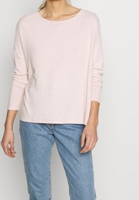 Haut rose pâle à manches longues avec un large col, fabriqué à partir d'un matériau doux, associé à un jean en denim bleu clair. Design simple et coupe décontractée.