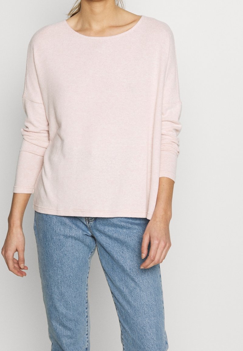 Haut rose pâle à manches longues avec un large col, fabriqué à partir d'un matériau doux, associé à un jean en denim bleu clair. Design simple et coupe décontractée.