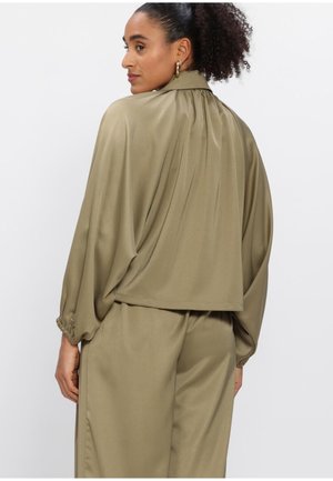 Vrouw met krullend haar, gekleed in een losse, olijfgroene satijnen blouse en bijpassende broek, die iets achterover gericht staat tegen een effen achtergrond.