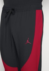 Pantalones deportivos negros y rojos con cintura elástica, cordón y un pequeño logo rojo de Jumpman en el muslo izquierdo. Tejido suave con un diseño deportivo.
