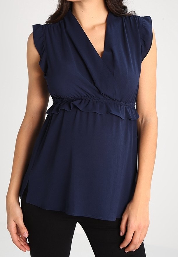 Mujer con blusa azul marino sin mangas, escote en V profundo y detalles de volantes en la cintura, combinada con pantalones negros.