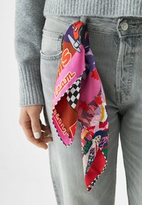 Écharpe en soie multicolore avec des graphismes vibrants, incluant des motifs et du texte, nouée à la poche d'un jean gris clair.