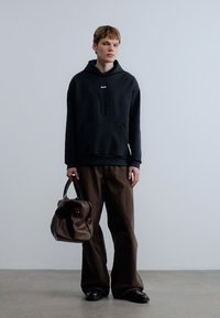 Μαύρο φούτερ με κουκούλα, μπροστινή τσέπη και κορδόνια, συνδυασμένο με άνετο καφέ παντελόνι. Αξεσουάρ περιλαμβάνει καφέ δερμάτινη τσάντα, στέκεται πάνω σε απλό φόντο.