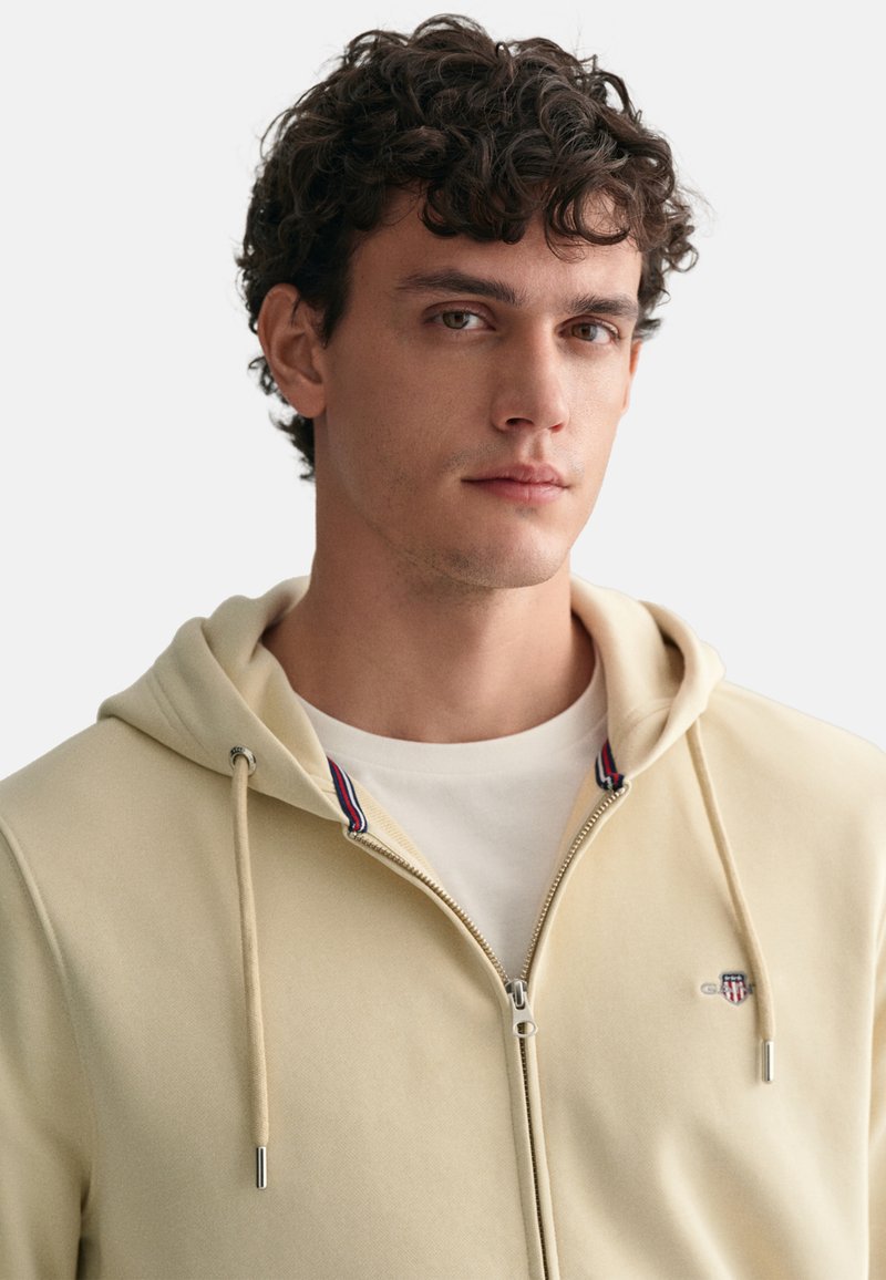 Beige Zip-Hoodie mit Kordelzügen, aus einem texturierten Stoff, einem weißen Innenshirt und einem kleinen Logo auf der Brust. Rote und blaue Akzente am Kragen.