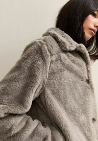 Gray faux fur coat with a hood, featuring a textured finish and button closures. The fabric appears soft and plush with a relaxed fit.