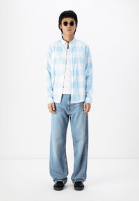 Hellblau kariertes Kurzarmhemd, über einem weißen T-Shirt getragen; kombiniert mit lockeren hellblauen Jeans und schwarzen Turnschuhen.