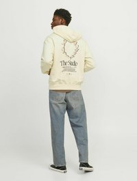 Beige hættetrøje med grafisk print på ryggen, sat sammen med lyseblå jeans og sorte sneakers. Hættetrøjen har snoretræk og en afslappet pasform.