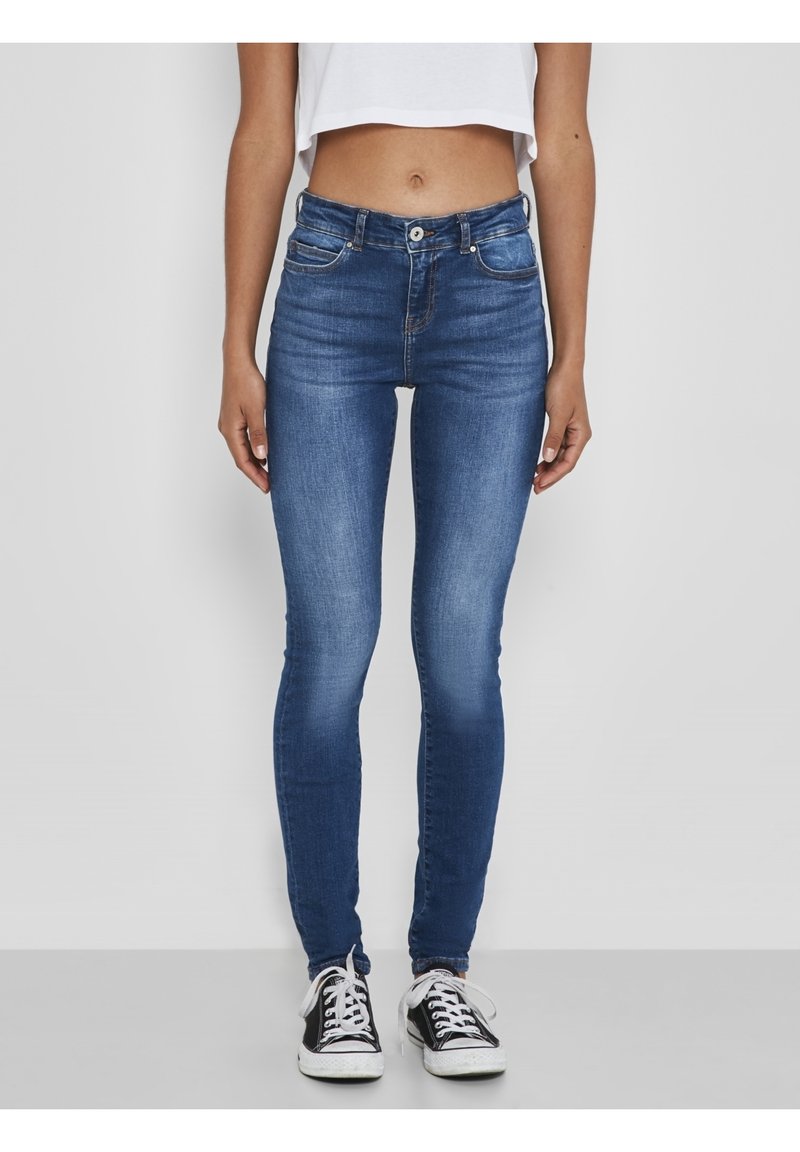 Jeans ajustados azules con una textura suave, que cuentan con bolsillos delanteros, cierre de botón y una cintura de altura media. Llevados con zapatillas negras.