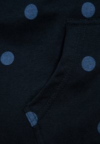 Tessuto nero con pois azzurri, dotato di una tasca con motivo a pois abbinato. Texture liscia e dettagli di cucitura curvi.