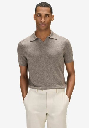 Man in een taupekleurig gebreid polohemd met korte mouwen, ingestopt in een hoge taille crème broek, met handen in de zakken, staande tegen een witte achtergrond.