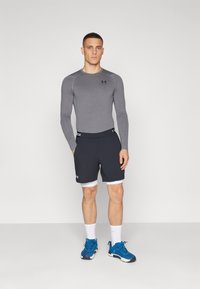 Graues langärmliges Sportoberteil mit Stretch, kombiniert mit blauen Shorts mit weißem Saum. Blaue Sportschuhe und weiße Socken runden den Look ab.