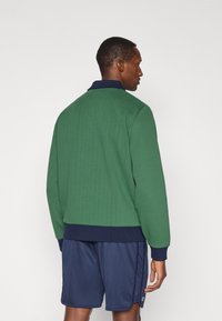Sweatshirt verde com textura e com colarinho e punhos em azul-marinho, combinado com calções azul-marinho. O design apresenta um corte descontraído e um tecido texturizado.