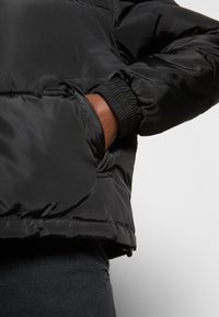Giacca nera imbottita con polsini elasticizzati, tasche con zip e un esterno liscio e lucido. L'orlo presenta un sistema di regolazione con cordino.