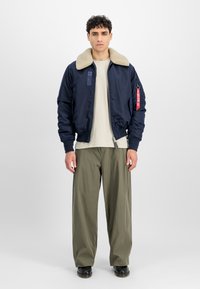 Marine bomberjakke med en beige shearlingkrave, lynlåslukning og burgunderetiket. Sammen med en cremefarvet skjorte og olivengrønne, vide bukser.