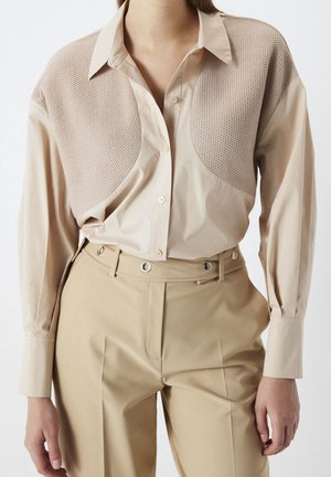 Chemise beige à manches longues avec panneaux texturés aux épaules associée à un pantalon kaki clair taille haute avec passants pour ceinture et œillets métalliques.