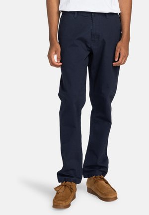 Navy blue cotton trousers with a straight fit, front pockets, and a zipper fly, paired with brown suede moccasin shoes.