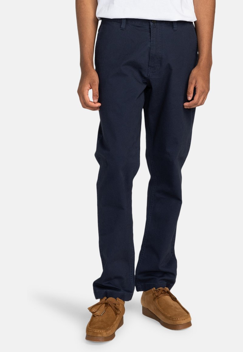Navy blue cotton trousers with a straight fit, front pockets, and a zipper fly, paired with brown suede moccasin shoes.