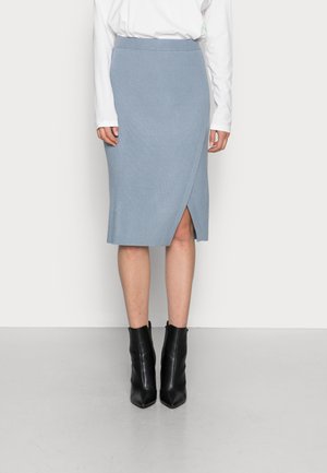 Kobieta ubrana w jasnoniebieską prążkowaną spódnicę do kolan z przednim rozcięciem, białą bluzkę z długim rękawem oraz czarne, ostro zakończone botki, stojąca na szarej podłodze.