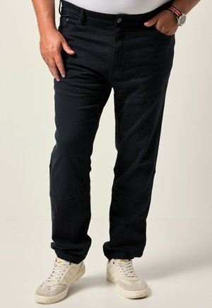 Man wearing black tapered pants with elastic cuffs, white sneakers, white shirt, and wristwatch with Norwegian flag strap.