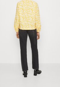 Femme portant un chemisier à manches longues jaune à motifs floraux, un jean noir droit et des bottines à talons noires, debout face à un mur blanc.