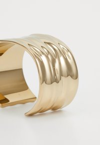 Gullfarget mansjettarmbånd med en bølgete design, glatt tekstur og åpen kutt. Overflaten reflekterer lys og forsterker det flytende mønsteret.