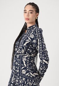 Abito blu navy con motivi floreali e geometrici, caratterizzato da un colletto e maniche lunghe, stretto in vita con elastico.