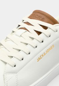 Bijele kožne tenisice s ravnim bež vezicama, smešnim kožnim detaljem na jeziku i urezanim zlatnim logotipom "JACK&JONES" sa strane.