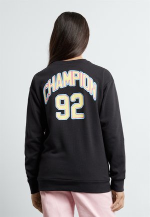 Person med langt mørkt hår iført sort sweatshirt med farverig "CHAMPION 92"-tekst på ryggen og lyserøde bukser, vendt væk fra kameraet.