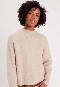 Pull en maille beige avec un col montant. Texture douce, coupe décontractée et poignets côtelés. Variations de couleur subtiles à travers le tissu.