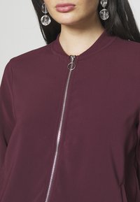 Burgundská bunda na zip s kulatým límcem, sázena na stříbrný zip a měkkou, hladkou texturu; doplněná o průhledné kulaté náušnice.