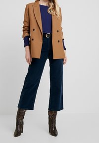 Blazer marrón de doble botonadura sobre un suéter azul marino, combinado con pantalones de pana anchos de color azul oscuro y tacones altos de estampado de serpiente marrón.