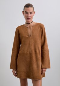 Brun ruskinds kjole med lange ærmer, en dyb halsudskæring og en løs pasform. Fremhævende sømme og en buet kant tilføjer detaljer til designet.