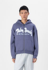 Lilla zip-up hættejakke med frontlommer, der har "Palm Beach Florida" i hvid skrift over brystet. Blødt, tekstureret stof.
