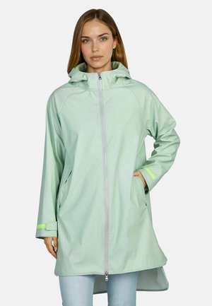 Junge Frau trägt eine hellgrüne Regenjacke mit Kapuze, Reißverschluss vorne und Seitentaschen, steht mit einer Hand in der Tasche.