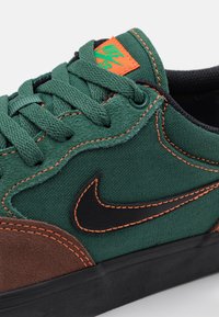Green fabric sneaker with brown suede accents, black Nike logo, orange stitching, and a padded collar. Features flat laces and metal eyelets.