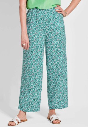 Grüne, blumengemusterte Hose aus leichtem Stoff, mit einem elastischen Bund und weitem Bein. Dazu werden weiße Sandalen getragen.