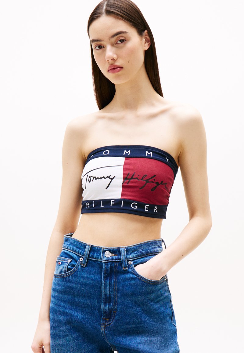 Mouwloze crop top in marineblauw, wit en bordeauxrood met logo-tekst, met een aansluitend ontwerp. Gepaarde met hoge taille blauwe jeans.
