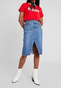 Falda lápiz de mezclilla en azul claro con una abertura frontal, dobladillo deshilachado y un diseño de cintura alta; combinada con una camiseta gráfica roja y botas blancas de tacón.