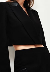 Blazer noir cropped avec accents brillants noirs sur les épaules, en tissu doux, présentant une coupe ajustée et un style ouvert à l'avant.